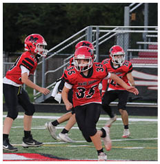 Two people wearing red football uniforms and helmets are engaged in a game on a field with a fence and bleachers in the background. Description generated by AI