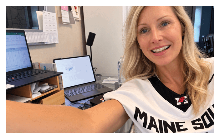 A person with long hair wearing a sports jersey is smiling and taking a selfie in an office setting with laptops and papers visible in the background. Description generated by AI