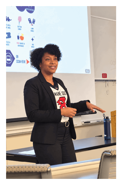 A person with curly hair wearing a blazer and a white shirt is smiling and looking slightly to the side in front of a blank white screen. Description generated by AI