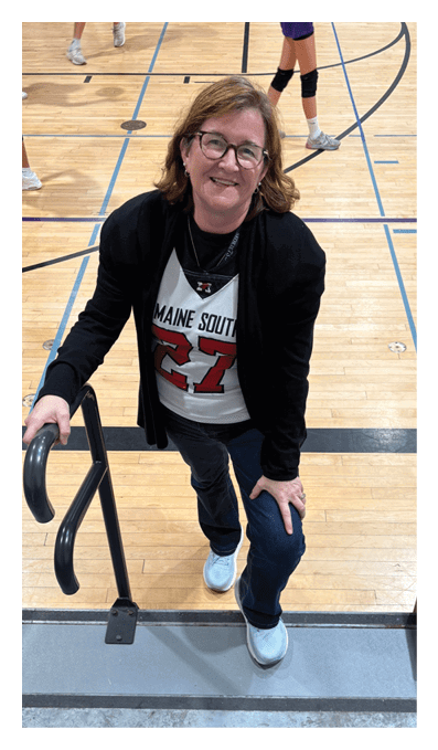 A person wearing glasses, a black jacket, and a sports jersey with the number 27 is smiling while standing on gym bleachers and holding a handrail, with a basketball court and players visible in the background. Description generated by AI