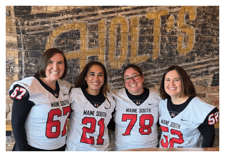 Four people wearing matching sports jerseys with numbers stand smiling in front of a brick wall with a large painted sign. Description generated by AI