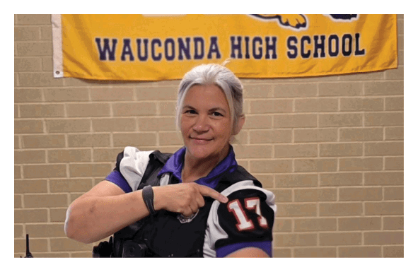 A person with light-colored hair is standing in front of a yellow banner with a large purple W, a bulldog mascot, and the text “Wauconda High School" on a brick wall background. Description generated by AI