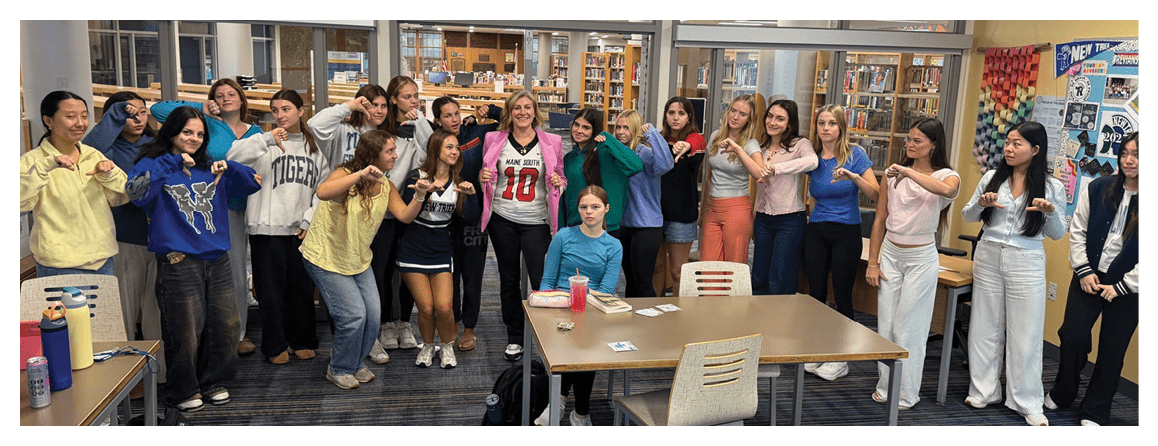 A group of people standing in a library are posing with one person sitting at a table in front of them while most of the standing people point towards the seated person. Description generated by AI