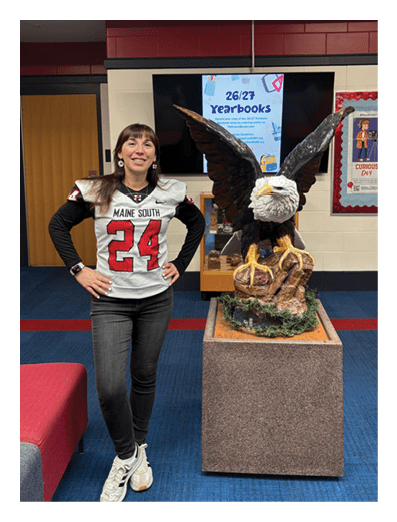A person wearing a sports jersey with the number 24 stands next to a large eagle statue inside a school hallway with yearbook posters in the background. Description generated by AI