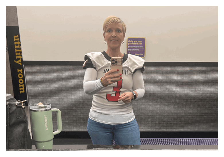 A person wearing a sports jersey and jeans is taking a mirror selfie in a restroom with a green insulated cup and a black bag on the counter. Description generated by AI