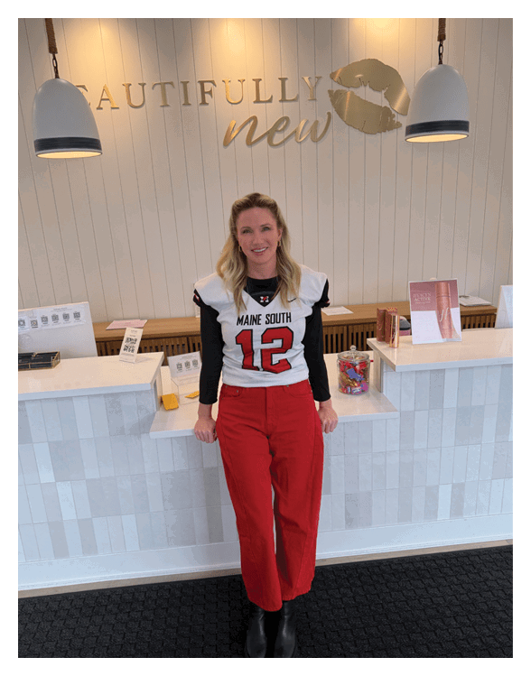 A person with long hair wearing a sports jersey that says “Maine South" is smiling and standing in front of a wall with the words "utifully new" and a large gold lip print. Description generated by AI