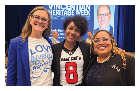 Three adults smiling and standing close together at an event with a backdrop reading “Vincentian Heritage Week" where one person wears a shirt with the phrase "Love is Inventing to Infinity," another wears a sports jersey with the number 8, and the third wears a dark blazer with embroidered initials. Description generated by AI