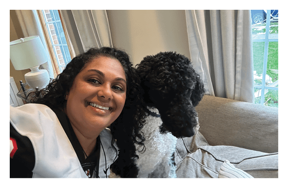 A person with curly hair wearing a white and black sports jersey is smiling and sitting next to a black and white curly-haired dog on a couch near a window with curtains. Description generated by AI