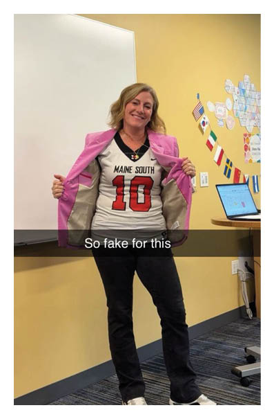 A person wearing a pink jacket over a sports jersey with the text “MAINE SOUTH 10" is standing in a classroom with a whiteboard and a laptop on a desk, smiling and holding the jacket open. Description generated by AI