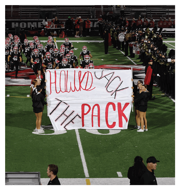 Four people in black outfits hold a large white banner with red and black letters reading “Hawks Pack The Pack" on a football field as a team in black uniforms and red helmets runs behind them and a marching band stands on the sidelines. Description generated by AI