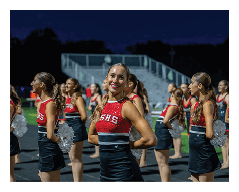 A group of adults in red and black cheerleading uniforms stand in formation on a sports field at night, with one person smiling and facing the camera. Description generated by AI