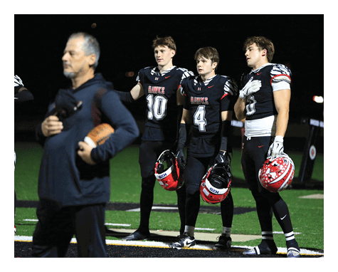 Three people in football uniforms holding helmets stand on a field while another person in casual clothing holds a football in the foreground. Description generated by AI