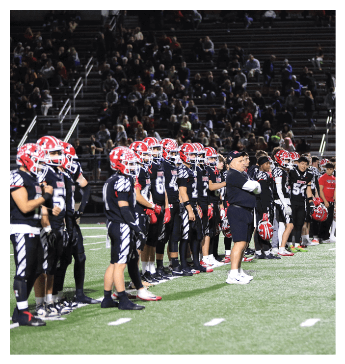 A group of football players in black uniforms with red helmets and a coach in black sportswear stand in a line on a football field with spectators in the background. Description generated by AI
