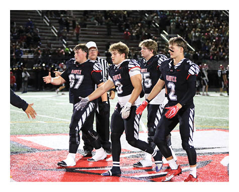 Four people in black football uniforms walk together on a field with a large red and white logo while a crowd watches from the stands. Description generated by AI