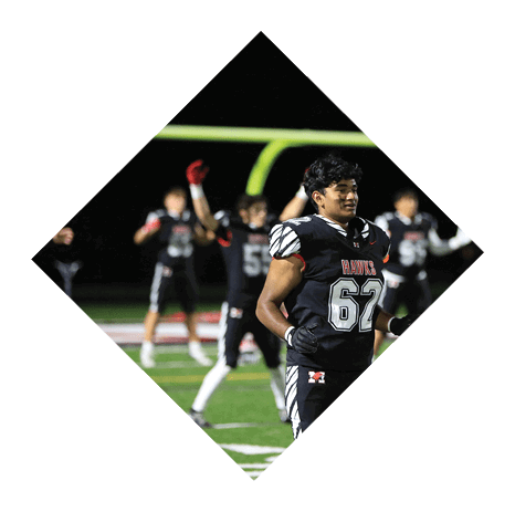 A person wearing a black football uniform with the number 62 stands on a field at night while other players in similar uniforms are visible in the background near a goalpost. Description generated by AI