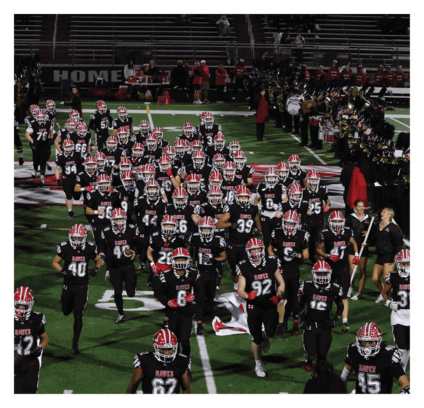 A group of people in black football uniforms with red and white helmets are walking onto a field while a marching band in black uniforms with tall hats plays instruments on the sidelines. Description generated by AI