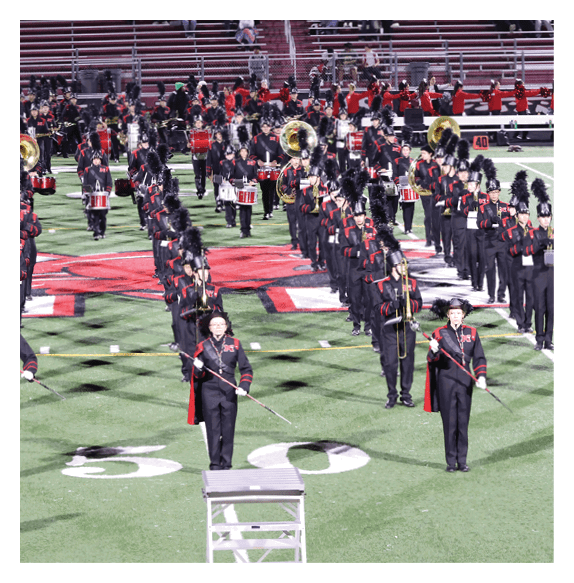 A marching band dressed in black uniforms with tall plumed hats is performing on a football field with red bleachers in the background. Description generated by AI