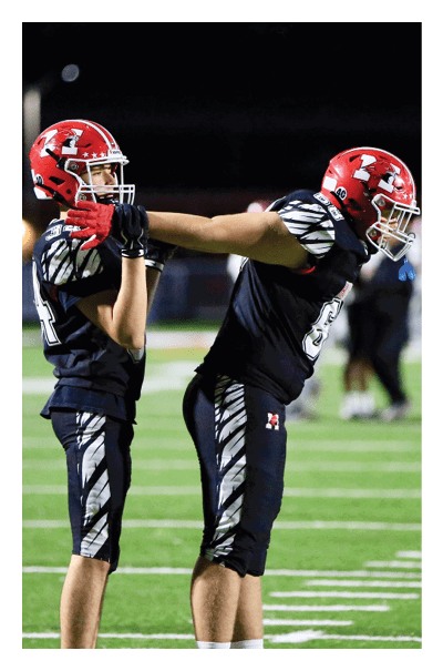 Two people wearing football uniforms with red helmets are standing on a football field at night, with one person holding the other's face mask. Description generated by AI