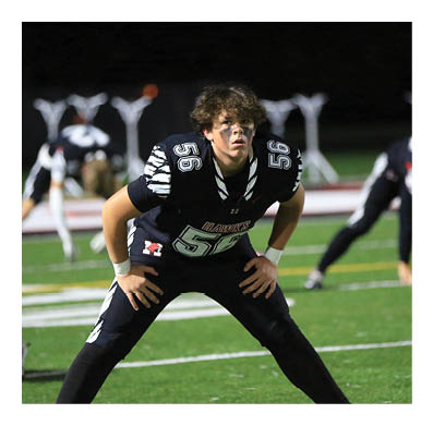 A person in a black football uniform with the number 56 is in a ready stance on a green field at night with other players and helmets visible in the background. Description generated by AI
