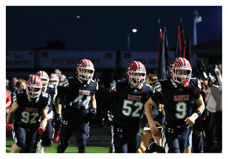 Several people in football uniforms with helmets are running onto a field at night with spectators and photographers in the background. Description generated by AI