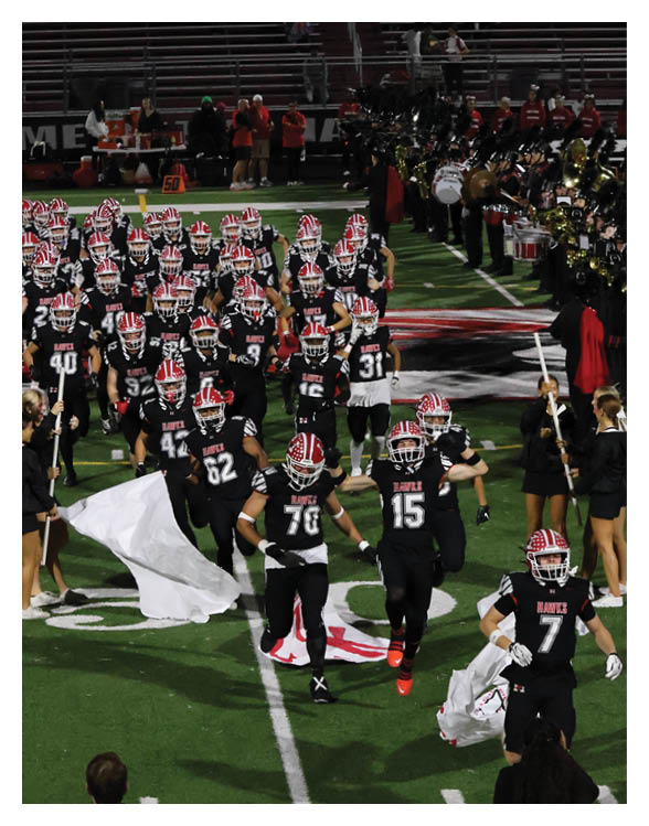 People in football uniforms are running onto a field from the left side while a marching band in black uniforms with tall black plumes stands in formation on the right side of the field. Description generated by AI