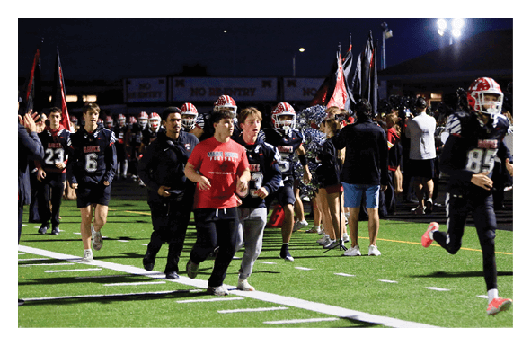 People in football uniforms and casual clothing are running onto a lit football field at night with cheerleaders and flags in the background. Description generated by AI