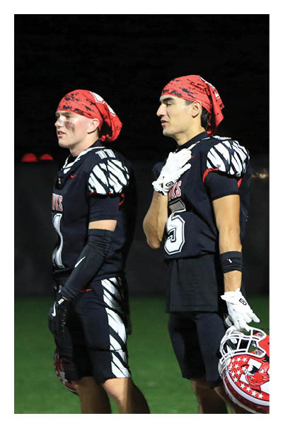 Two people wearing red bandanas with black patterns are shown against a dark background, with one person facing sideways and the other partially visible. Description generated by AI