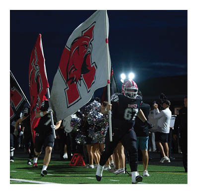 People in sports uniforms run on a field at night holding large flags with a red and black logo while others stand in the background near bright lights. Description generated by AI