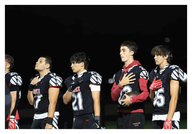 Five people in football uniforms stand side by side on a field at night with their right hands over their hearts. Description generated by AI