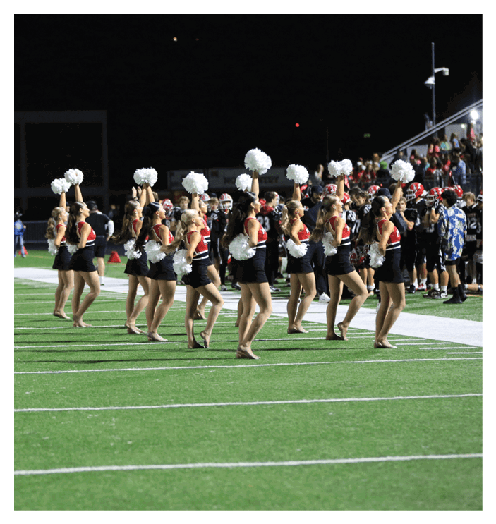 A group of people dressed in coordinated outfits with pom-poms are performing a cheer or dance routine on a football field at night. Description generated by AI