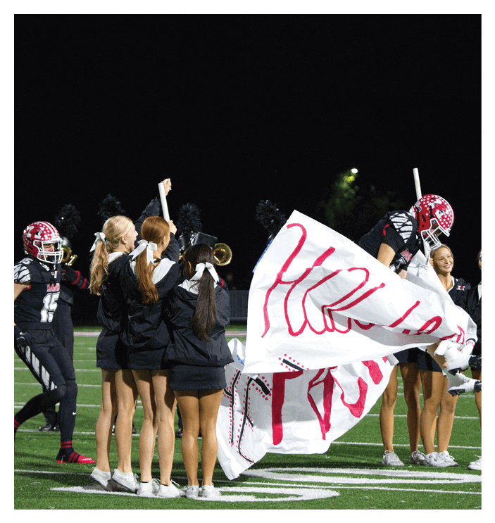 People in sports uniforms and cheer outfits are preparing to break through a large banner on a football field at night. Description generated by AI
