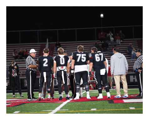 Four people in football uniforms and two adults stand on a football field at night near the center logo while a referee talks to them. Description generated by AI
