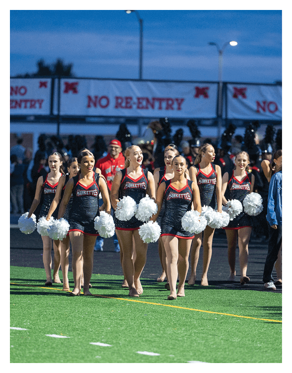A group of adults in matching cheerleading uniforms holding white pom-poms are walking on a sports field with a “No Re-Entry" sign visible in the background. Description generated by AI
