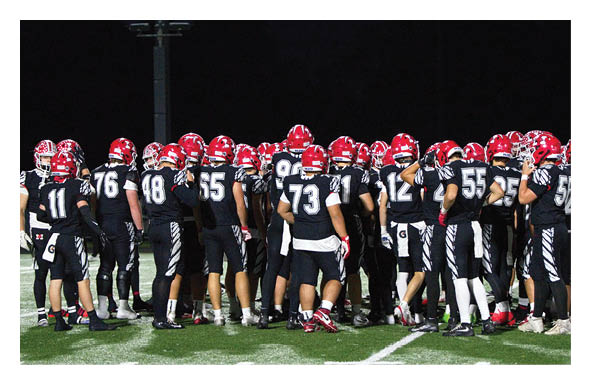 A group of people wearing black sports uniforms and red helmets are gathered closely together on a football field at night. Description generated by AI