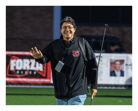 A person wearing glasses and a black jacket with a football logo is smiling and waving while standing outdoors at night. Description generated by AI
