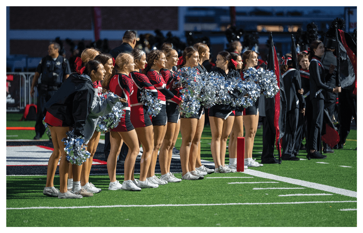 A group of people in matching athletic uniforms stand on a sports field holding pom-poms while others in band uniforms stand nearby. Description generated by AI