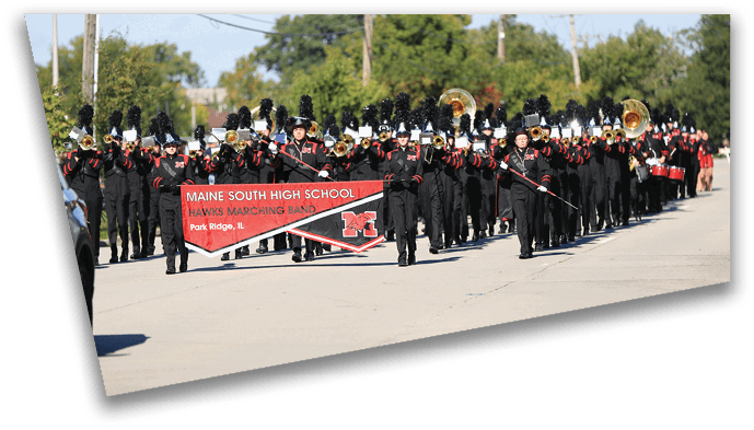 A marching band dressed in black uniforms with tall black plumes is performing outdoors on a street, holding a red banner that reads Maine South High School Hawks Marching Band. Description generated by AI