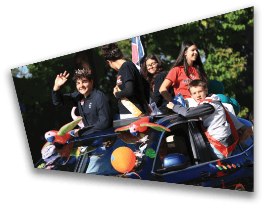 People wearing casual clothing are riding on and standing around a decorated dark blue vehicle with colorful balloons and parrot decorations in an outdoor setting. Description generated by AI