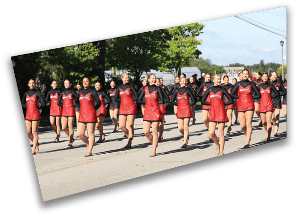 A group of people dressed in matching red and black outfits are marching or dancing in formation outdoors on a sunny day. Description generated by AI