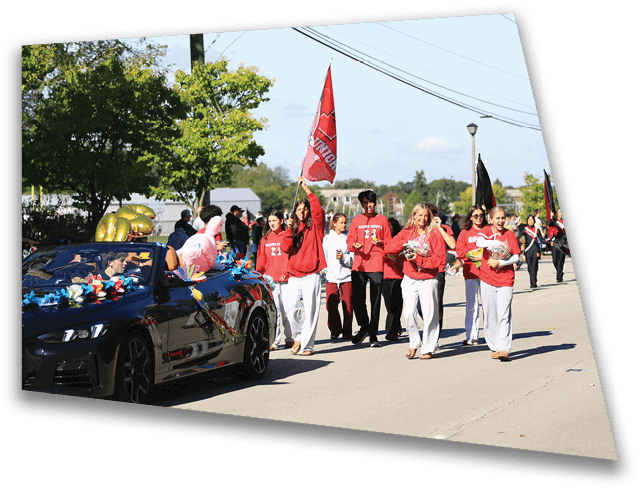 A group of people wearing red and white clothing are walking in a parade behind a black car decorated with colorful ribbons and balloons on a sunny day. Description generated by AI