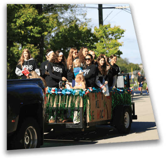A group of people wearing matching black hoodies are riding on a decorated trailer attached to a vehicle during a sunny outdoor event. Description generated by AI