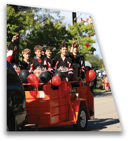 A group of people wearing matching sports jerseys are riding in a red trailer decorated with black and red balloons being towed by a black vehicle on a sunny street. Description generated by AI