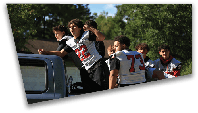 A group of people wearing sports jerseys are gathered outdoors near a vehicle, with some leaning on it and others standing close by. Description generated by AI