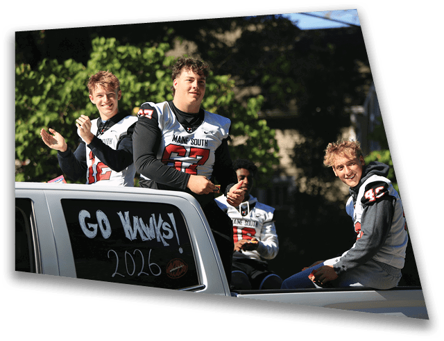Four people wearing sports jerseys are sitting and clapping on the back of a vehicle with a window sign that says “GO KAWKS! 2026" during a sunny outdoor event. Description generated by AI