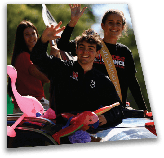 Three smiling people wave from a decorated car during a daytime outdoor event, with one wearing a sash that reads “Homecoming Royalty." Description generated by AI