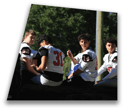 Three people wearing football jerseys with numbers 32, 31, and 14 are sitting on the back of a black vehicle outdoors with trees in the background. Description generated by AI