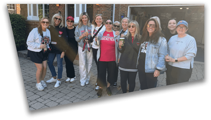 A group of adults standing outdoors on a paved area, smiling and casually dressed, with some holding drinks and wearing sunglasses. Description generated by AI