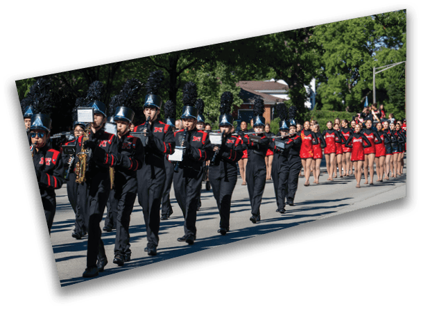 A marching band dressed in black uniforms with tall plumed hats is playing instruments while walking down a street followed by a group of people in red and black outfits. Description generated by AI