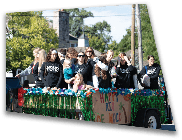 A group of people wearing matching black shirts with white text are standing on a decorated float with colorful garlands and a sign that reads “Happy Homecoming." Description generated by AI