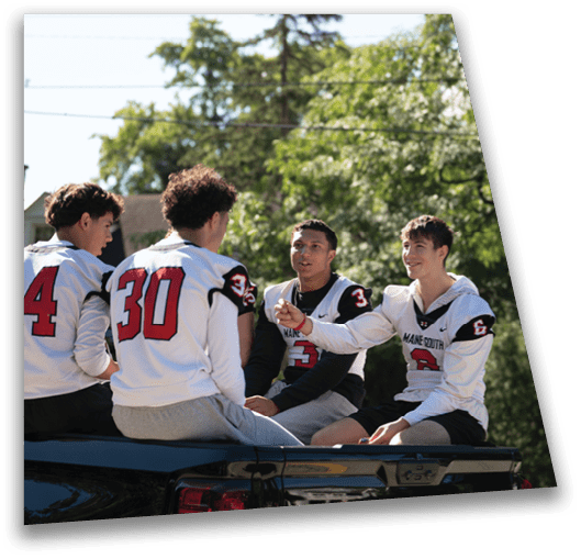 Three people wearing sports jerseys are sitting and talking on the back of a vehicle outdoors with trees in the background. Description generated by AI